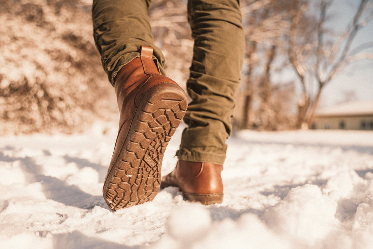 Eine Person in braunen Lederstiefeln und grünen Hosen läuft durch den Schnee. Die Sohlen der Stiefel haben ein strukturiertes Muster. Im Hintergrund sind kahle Winterbäume unter einem klaren blauen Himmel zu sehen, die eine ruhige Winterszene schaffen.