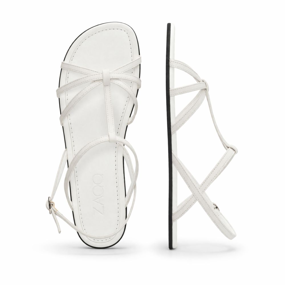 Wir präsentieren die QADA White Sandalen von ZAQQ, gefertigt aus hochwertigem Nappaleder. Diese eleganten Barfußsandalen haben dünne Lederriemen und eine glatte, flache Sohle. Eine Sandale ist von oben zu sehen, während die andere ihr schlankes Seitenprofil mit einer kleinen Knöchelriemenschnalle zeigt.