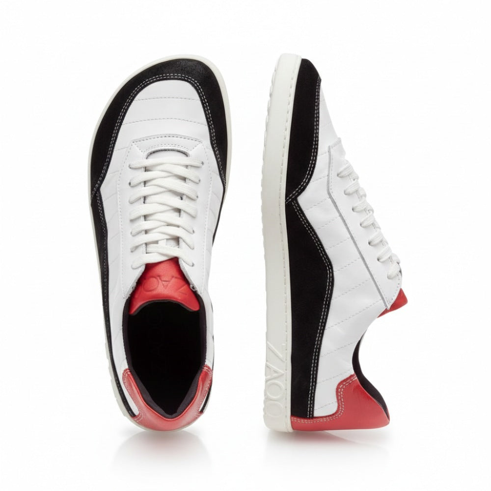 Ein Paar schwarz-weiße QAMPION-Sneaker von ZAQQ mit weißem Obermaterial, schwarzen Akzenten und roten Absätzen. Diese Schuhe zeichnen sich durch Velourslederdetails, weiße Schnürsenkel und eine strukturierte, graue, flexible Sohle aus. Ein Schuh steht aufrecht, während der andere auf der Seite liegt und den Markennamen ZAQQ deutlich auf der Seite der Sohle zeigt.