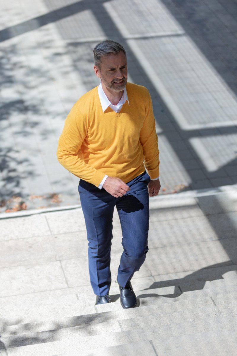 Ein Mann mit grauem Haar und Bart geht draußen eine Steintreppe hinauf. Er trägt einen leuchtend gelben Pullover über einem weißen Hemd mit Kragen, blaue Hosen und dunkle Schuhe. Schatten eines nahe gelegenen Gebäudes erzeugen Muster auf dem Boden und den Stufen.